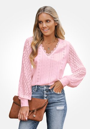 Vrouw met golvend blond haar die een roze kanten blouse en blauwe high-waisted jeans draagt, een bruine leren clutch vasthoudt, glimlacht en naar rechts kijkt.