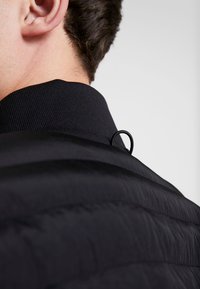 En närbild av kragen och ryggen på en mörk, puffy jacka, med en ögla för upphängning, som delvis avslöjar en persons nacke och öra.