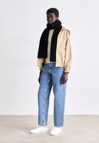 Giacca beige con zip e cappuccio, maglione beige, jeans blue a gamba dritta, sciarpa nera a coste e sneakers bianche. Texture dei tessuti lisci.