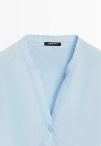 Chemise bleu clair sans col avec un seul bouton visible et une étiquette « Massimo Dutti » au niveau de l'encolure.