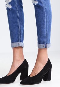Černé semišové lodičky na vysokém podpatku s špičatou špičkou a silným podpatkem, spárované s ohrnutými džíny z denimu s opotřebovanými detaily.