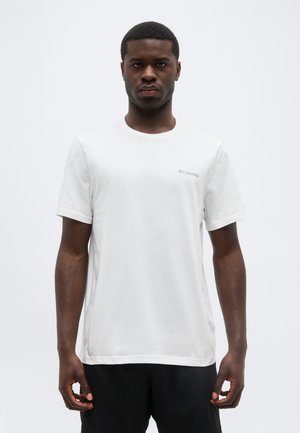 Άνδρας που φοράει απλό λευκό κοντομάνικο μπλουζάκι Columbia και μαύρο παντελόνι, στέκεται μπροστά σε απλό φωτεινό φόντο.