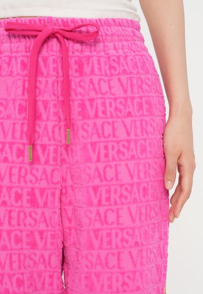 Vaaleanpunaiset froteehousut, joissa on kaikkialla embossattu "VERSACE" -kuvio, elastinen vyötärö, jossa on kiristysnauha, sekä kultaiset yksityiskohdat kärjissä.