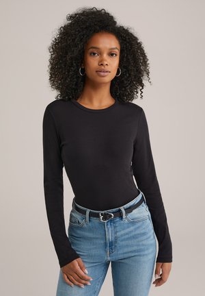 Vrouw met krullend haar, die grote oorbellen draagt, een zwart longsleeve shirt, lichtblauwe jeans en een zwarte riem, staande voor een effen achtergrond.
