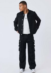 Giacca nera testurizzata con bottoni bianchi e pantaloni cargo abbinati con diverse tasche. Indossata sopra una maglietta bianca, abbinata a sneakers bianche.
