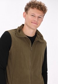 Olivgrüner Fleece-Weste mit Kragen und durchgehendem Reißverschluss, getragen über einem schwarzen Langarmshirt; hat eine glatte Textur und ein schlichtes Design.