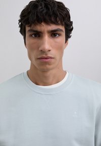 Joven con cabello rizado oscuro, vestido con un suéter azul claro de cuello redondo sobre una camisa blanca, frente a un fondo claro y liso.