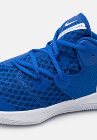 Modrá sportovní bota s horní částí z prodyšné síťoviny, úhlovým designem, výraznými šňůrkami a texturovanými akcenty. Obsahuje branding Nike a bílou gumovou podrážku.