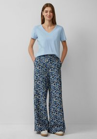 Lichtblauw V-hals T-shirt, gecombineerd met wijde broek met een bloemen-geometrisch patroon in blauw, crème en marineblauw. Witte sneakers.