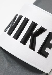 Balta šlepetė su tekstūruota juostele ir dideliu blizgiu juodu "NIKE" logotipu. Tamsiai pilkas padas papildo dizainą.