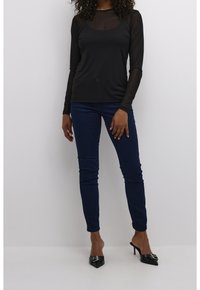 Haut noir à manches longues avec un overlay transparent, associé à un jean slim bleu marine et des escarpins noirs à bout pointu ornés d'accessoires décoratifs.