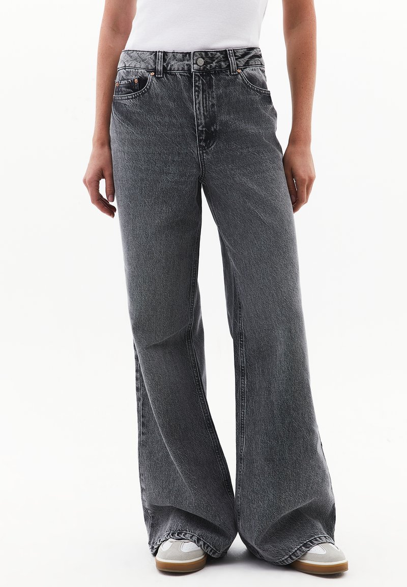 Jeans de pierna ancha en denim negro deslavado, con cintura alta, diseño de cinco bolsillos y detalles de costura sutiles. Combinados con zapatillas grises.