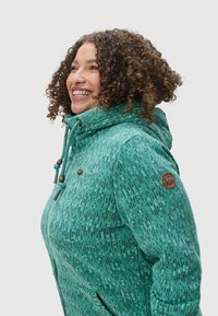 Veste teal à motifs avec capuche, fabriquée dans un tissu texturé, dotée d'un cordon de serrage et d'accents métalliques, ainsi que d'un badge logo sur la manche.