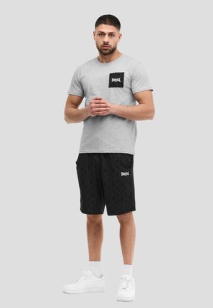 Uomo che indossa una maglietta grigia con logo nero sulla tasca, pantaloncini neri con motivo e logo bianco, calzini bianchi e scarpe da ginnastica bianche, in piedi contro uno sfondo grigio.