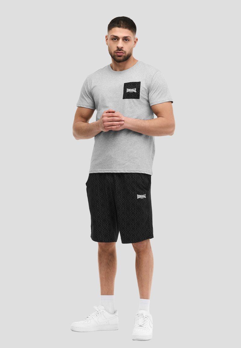 Man draagt een grijs T-shirt met een zwart logo op de borstzak, zwarte korte broek met patroon en wit logo, witte sokken en witte sneakers, staand tegen een grijze achtergrond.