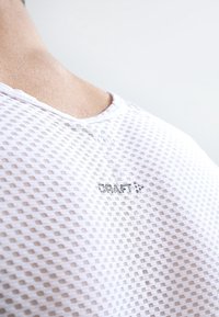 Maglietta bianca in mesh traspirante con scollo rotondo, caratterizzata da un piccolo logo "CRAFT" ricamato sul retro. Motivo testurizzato con lavorazione a jersey aperto.