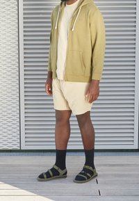 Hombre vestido con pantalones cortos beige, camiseta crema, sudadera con cremallera verde oliva, calcetines negros y sandalias verdes oliva, de pie sobre una plataforma de madera con un muro louvered detrás.