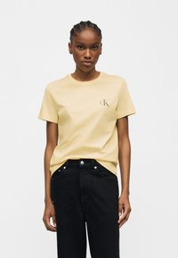 Γυναίκα με πλεγμένα μαλλιά που φοράει ένα ανοιχτό κίτρινο t-shirt Calvin Klein και μαύρο τζιν ψηλόμεσο σε μονόχρωμο φόντο.