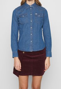 Jeansskjorta med spetskrage, långa ärmar och två bröstfickor; har silverfärgade tryckknappar och kontrasterande orange sömmar.