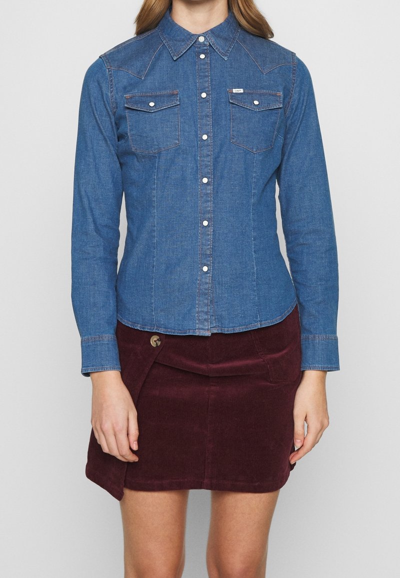 Jeansskjorta med spetskrage, långa ärmar och två bröstfickor; har silverfärgade tryckknappar och kontrasterande orange sömmar.