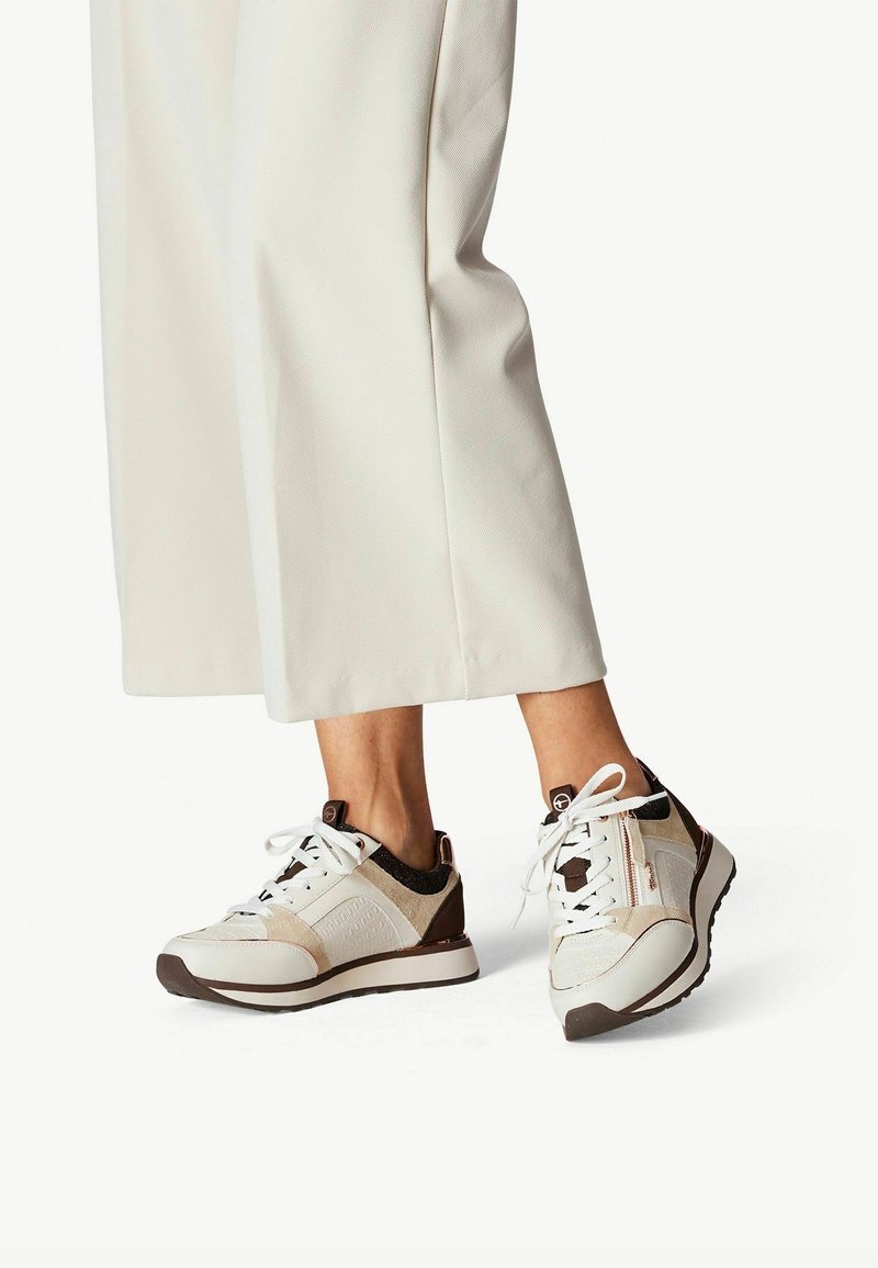 Bílé a béžové tenisky s texturovanými panely, hnědými akcenty a tkaničkami. Design obsahuje polstrovanou podešvi a detaily se zipem.