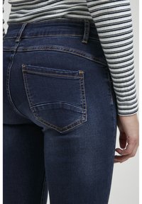 Jeans de mezclilla en azul oscuro, con un diseño ajustado, costuras visibles en un contraste claro y un bolsillo trasero con una forma angular.