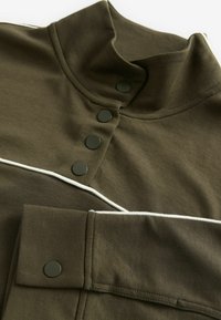 Haut en tricot vert avec un col montant, présentant des boutons-pression noirs et des accents de piping blanc le long du col et des manches. Texture douce.