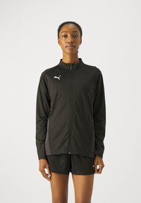 Svart zip-up jakke laget av lett stoff, med krage, sidelommer og en kontrasterende logo på brystet.