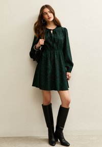 Groene luipaardprintjurk met een striksluiting, lange mouwen en een getailleerde taille. Zwarte kniehoge laarzen met hak maken de outfit compleet.