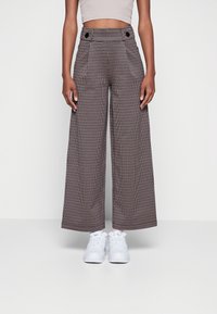 Kalhoty s širokými nohavicemi v hnědo-krémovém vzoru houndstooth, s vysokým pasem a dvojitými knoflíkovými detaily, v kombinaci s bílými teniskami.
