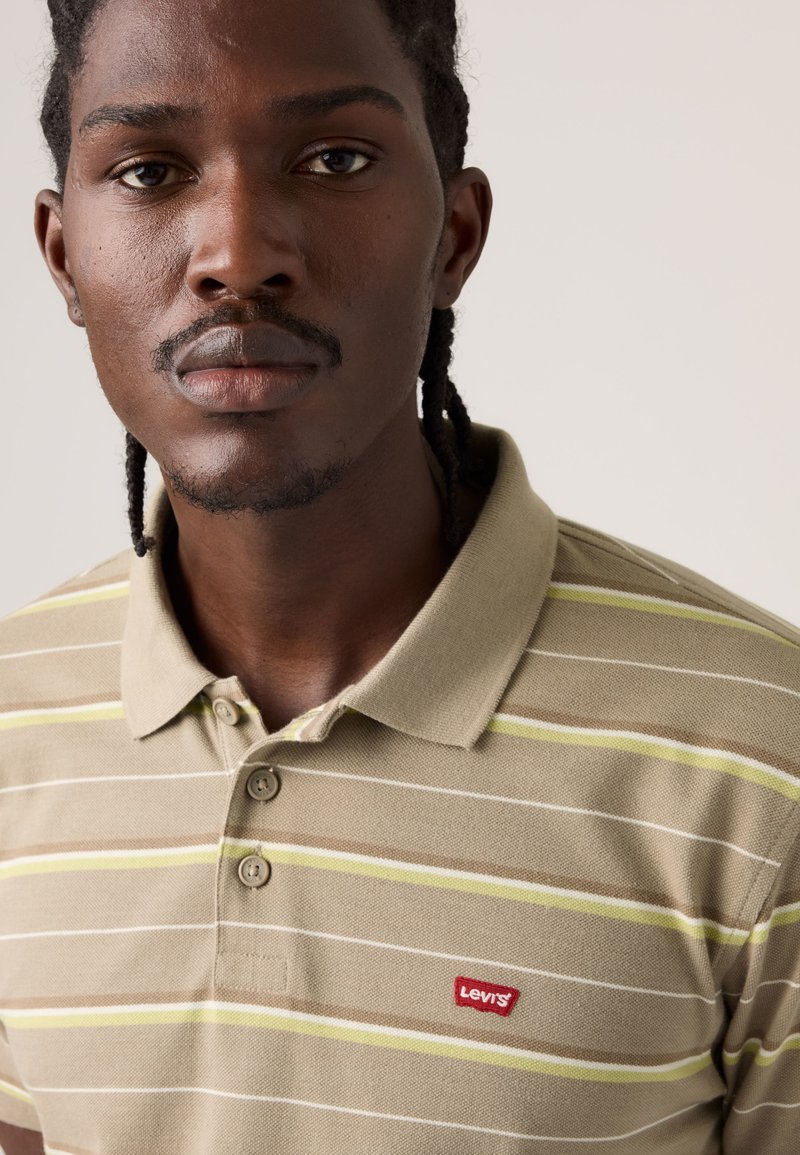 Young man with braided hair wearing a beige striped Levi’s polo shirt, looking directly at the camera against a plain background.