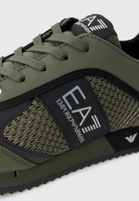 Grønn atletisk joggesko med mesh-paneler og en svart logo-patch. Teksturert såle med zigzag-mønster og flate snører.