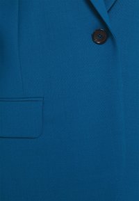 Niebieska marynarka z teksturowanej tkaniny, pojedynczym czarnym guzikiem i kieszonką na piersi. Dopasowany krój z gładkim wykończeniem.