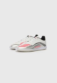 Witte sportieve schoenen met zilveren accenten, zwarte hiel en rode en zwarte strepen. Voorzien van een gestructureerde zool en "MESSi" branding op de zijkant.