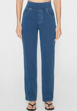 Frau trägt hoch taillierte, blaue, gerade geschnittene Jeans mit Vordertaschen und elastischem Bund, kombiniert mit schwarzen Flip-Flop-Sandalen.