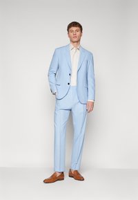 Suit azzurro chiaro con un design strutturato, caratterizzato da una giacca a un petto, pantaloni frontali piatti e una camicia crema. Mocassini in pelle marrone.