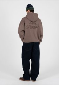Junger Mann, der einen braunen Hoodie mit dem Aufdruck "Prohibited 10119 Berlin", dunkle weite Jeans, braune Schuhe und eine schwarze Mütze trägt und mit dem Rücken zur Kamera steht.