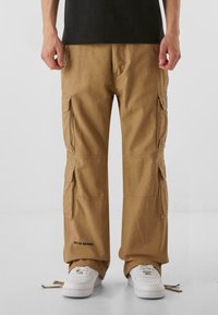 Ljusbruna cargobyxor i lätt tyg med flera sidofickor och elastiska muddar; har logodetaljer nära fållen. Kombinerat med vita sneakers.