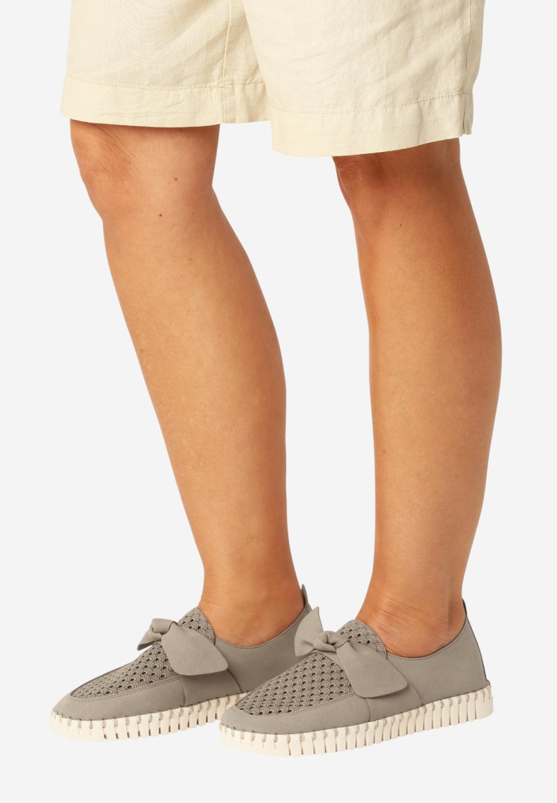 Beine, die beige Shorts und graue Slipper mit strukturiertem Obermaterial und dekorativer Schleife vor weißem Hintergrund tragen.