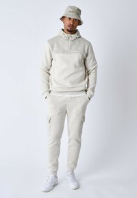 Sweatshirt blanc avec un empiècement gris à motifs, col zippé, et un pantalon de survêtement assorti doté de poches latérales et de motifs similaires, associé à des baskets blanches.