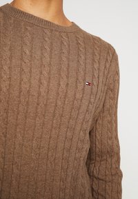 Pull en mailles torsadées marron avec un col rond et un petit logo brodé avec des accents rouge, blanc et bleu sur la poitrine.