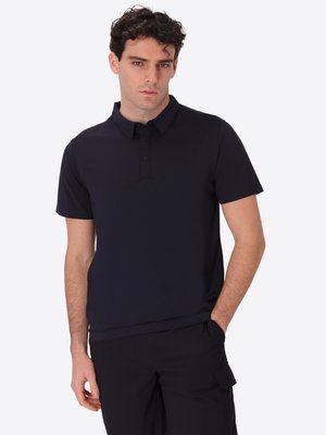 Giovane uomo con capelli ricci e scuri che indossa una polo nera a maniche corte e pantaloni neri, in piedi con una mano in tasca davanti a uno sfondo bianco.