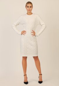 Weißes Strickkleid mit Kabeldesign, langen Ärmeln, geripptem Saum und geradem Schnitt. Kombiniert mit schwarzen Riemchen-Absätzen.