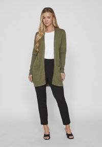 Olive green long cardigan with open front, ribbed hem, and two front pockets, paired with a white top and black fitted pants.