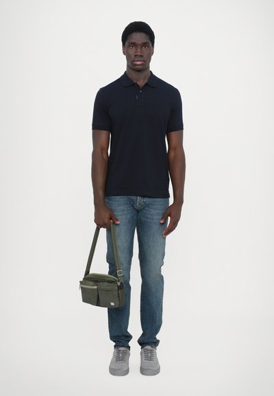 Navy polo shirt, light blue jeans, gray sneakers, holding a green messenger bag with two front pockets and a shoulder strap.