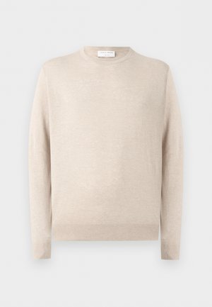 Göndör hajú férfi bézs színű hosszú ujjú ingben, fekete nadrágban és fekete cipőben, áll és oldalra néz fehér háttér előtt.