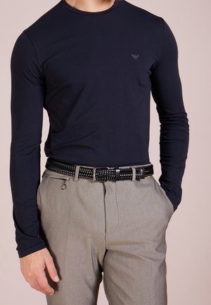 Man wearing dark navy long-sleeve shirt and light gray trousers with black woven belt, standing with one hand in pocket.