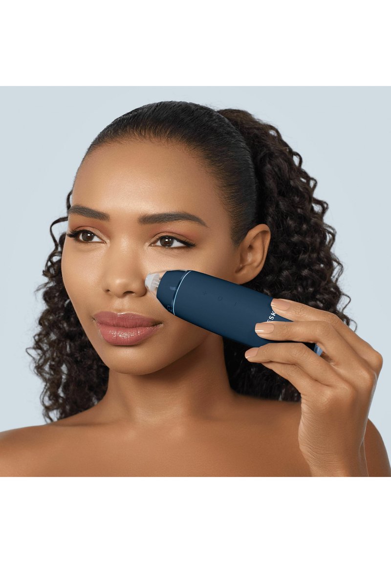 Woman using a dark blue facial device on her nose, focusing on skincare, with curly hair pulled back and neutral background.