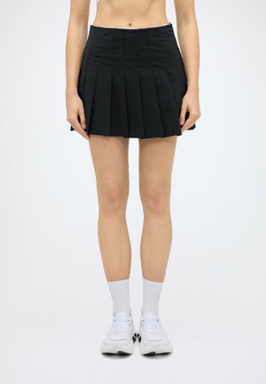 Femme portant une jupe plissée noire, des chaussettes blanches et des baskets blanches, debout devant un fond clair et uni.