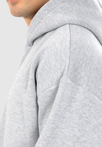 Haut à capuche gris clair avec une coupe décontractée, un tissu texturé doux et un capuchon avec cordon de serrage. Logo brodé discret sur la zone de la poitrine.